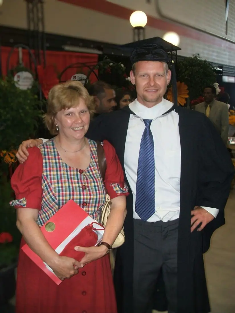 A man in a graduation cap and gown standing with his arm around a smiling woman in a red dress who holds a red diploma folder