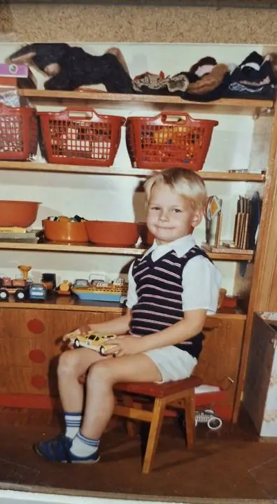 A blond little boy in a striped vest sitting on a wooden stool holding a toy car in front of a shelf of toys
