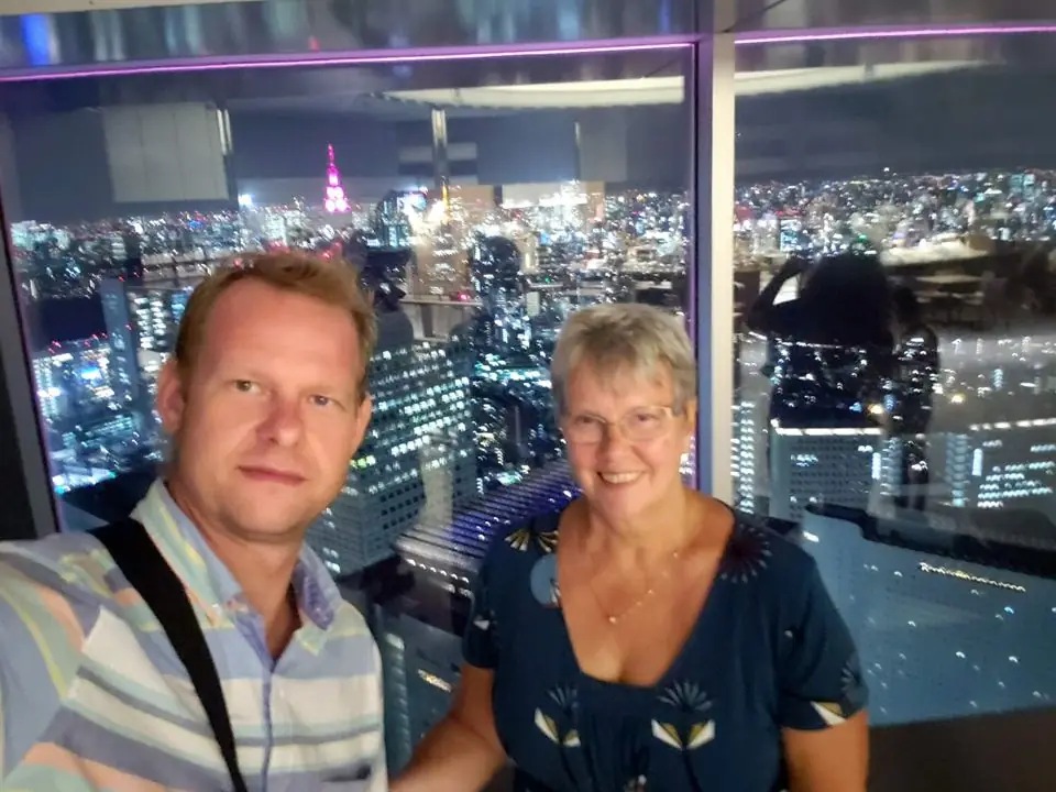 A man and his mother taking a selfie in front of a window with the Tokyo skyline glittering at night behind them