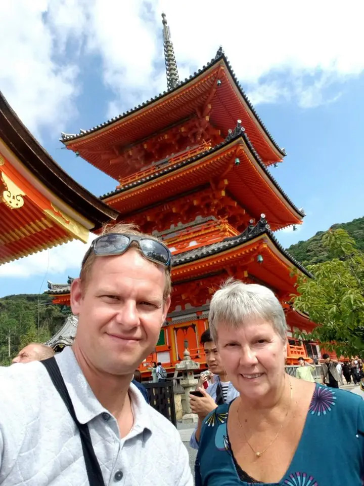 A smiling man with sunglasses on his head and his mother taking a selfie in front of a bright orange three-story pagoda in Kyoto