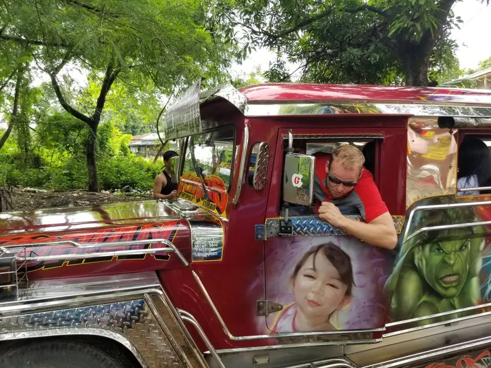A man in sunglasses leaning out the window of a brightly painted red jeepney decorated with airbrushed art in the Philippines