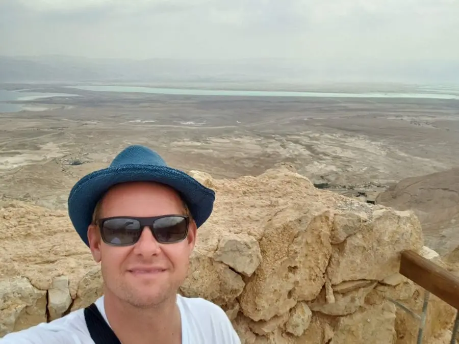 A man in a blue hat and sunglasses taking a selfie on a rocky mountaintop above a hazy desert and the Dead Sea