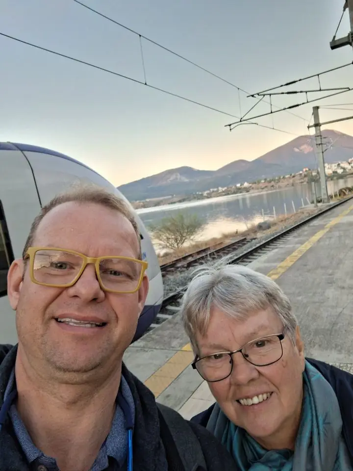 A man in yellow glasses and his mother taking a selfie on a train platform beside a lake, with mountains in the evening light behind them