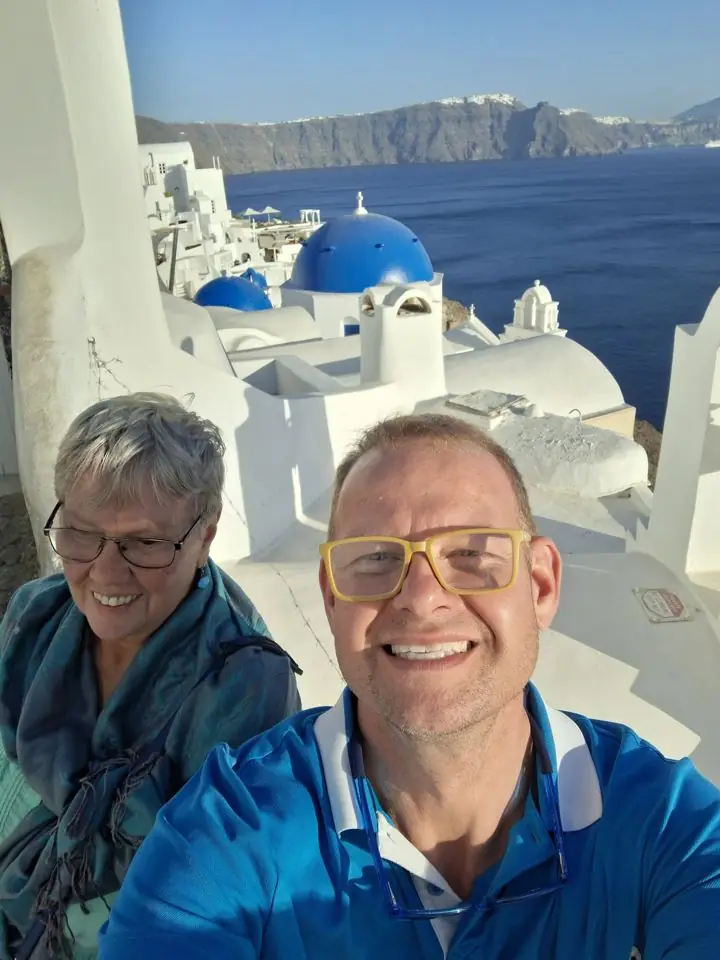 A man in yellow glasses and a blue shirt and his mother taking a selfie among whitewashed buildings and blue domes above the sea in Santorini