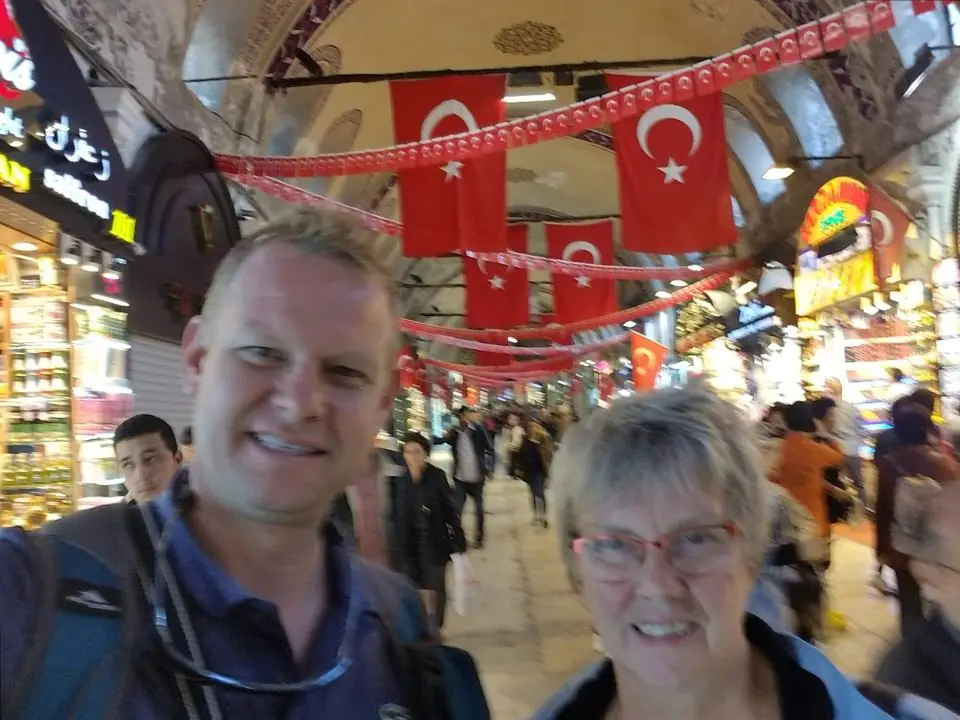 A smiling man and his mother taking a selfie in a busy covered bazaar in Istanbul, with rows of red Turkish flags hanging overhead